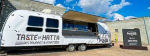 عربة طعام متخفية باللون الأبيض تحمل شعار Taste of Hatta تقدم الوجبات السريعة إلى الخارج في الهواء الطلق.