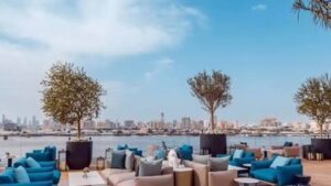 سطح خارجي فسيح يمثل مقاعد مريحة بدرجات اللون الأزرق الذهبي مع الزينة والنباتات المطلة على مياه خور دبي باللون الأزرق تحت سماء صافية.